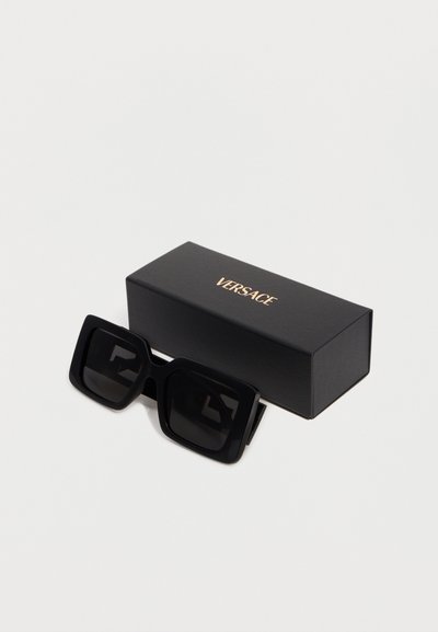 Μαύρα υπερμεγέθη τετράγωνα γυαλιά ηλίου που ακουμπούν δίπλα σε μια μαύρη ορθογώνια θήκη με το λογότυπο της Versace, σε λευκό φόντο.