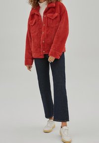 Rode jas met fleece voering, knoopsluiting aan de voorkant, twee borstzakken en een ontspannen pasvorm. Gecombineerd met donkerblauwe uitlopende jeans en witte sneakers.