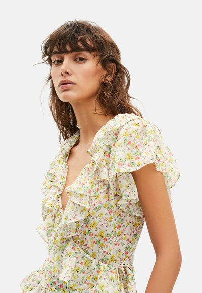 Joven mujer con cabello castaño ondulado que lleva un vestido floral claro con detalles con volantes y mangas cortas y onduladas, sobre un fondo liso.