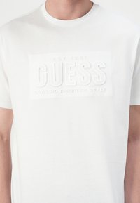 Hvit bomull t-skjorte med en teksturert, hevet hvit logo med "ETABLERT 1981" og "KLASSISK AMERIKANSK STIL" i fet skrift.