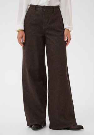 Person trägt eine hoch taillierte, weit geschnittene, dunkelbraune Hose und eine weiße, langärmelige Bluse, steht vor einem schlichten, hellen Hintergrund.