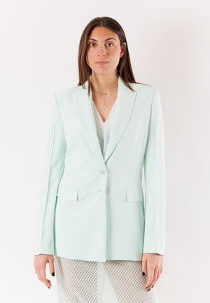 Donna con i capelli castani lunghi che indossa una giacca color menta chiaro sopra una maglietta bianca e una gonna bianca in rete su uno sfondo bianco semplice.