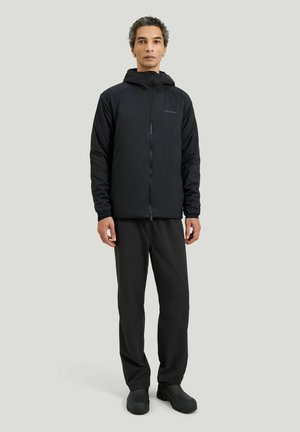 Mann steht und trägt eine schwarze Kapuzenjacke mit Reißverschluss, schwarze Hose und schwarze Schuhe vor neutralem Hintergrund.