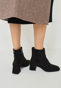 Black suede ankle boots with a square heel and side zippers, featuring a smooth texture and a minimalist design.