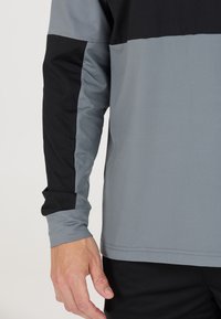 Tröja med långa ärmar för sportbruk som har en ljusgrå och svart färgblockdesign, med slät textur och justerbara ärmslut.