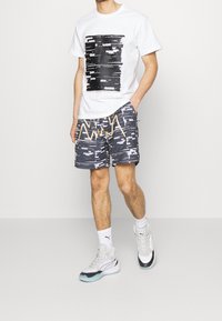 Hvid t-shirt med sort grafisk design, kombineret med marineblå mønstrede shorts med gyldne detaljer, og grå-hvide træningssko.