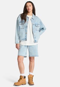 Veste en denim bleu clair avec deux poches poitrine et des boutons, associée à un short en denim assorti et des bottes beiges avec des lacets jaunes.