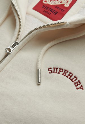 Cremefarvet Superdry sweatshirt med lynlås, delvist åbnet lynlås, rød broderet logo, snor med metaltip og rød mærkat indeni kraven.