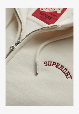 Sudadera de cremallera color crema de Superdry con cremallera parcialmente abierta, logotipo bordado en rojo, cordón con punta de metal y etiqueta roja dentro del cuello.