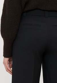 Black tailored trousers with a smooth texture, featuring two back pockets and a slim fit. The waistband is slightly elevated, ensuring a tailored look.