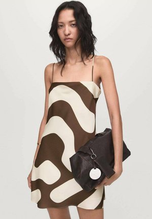 Mujer vestida con un vestido de tirantes de patrón abstracto en tonos marrón y crema, sosteniendo un gran bolso de mano oscuro marrón contra un fondo plano.