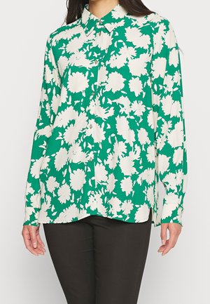 Femme portant un chemisier vert à boutons avec un grand motif floral blanc et un pantalon noir, debout les bras détendus le long du corps.