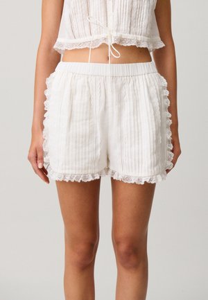 Pantalones cortos blancos de talle alto con banda elástica, rayas verticales finas y borde de encaje con ondulaciones, usados por una persona con las manos a los lados.