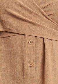 Svetlo rjavo tkano blago z mehkimi teksturami, ki ima prečnim dizajnom in gumbom, kar ustvarja nežno drapiranje.