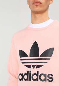 Man bär rosa sweatshirt med stor svart Adidas treklöverlogga och text på framsidan, vit tröja med rund hals under.