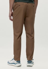 Brūni bikses ar gludu virsmu, sašaurinātu dizainu un divām aizmugures kabatām; apvienotas ar pelēkiem un baltajiem sporta apaviem ar zila akcentiem.