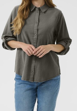 Femme portant une chemise boutonnée à manches longues vert olive avec manches retroussées et un jean bleu clair, les mains jointes de manière détendue devant elle.