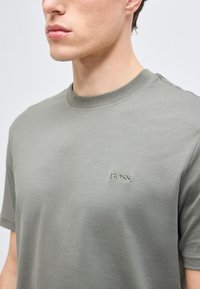 Νέος άνδρας που φοράει ανοιχτό γκρι μπλουζάκι με στρογγυλή λαιμόκοψη και μικρό κεντημένο λογότυπο "BOSS" στο αριστερό μέρος του στήθους.