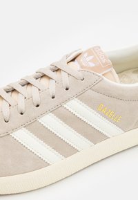 Beige suede sneaker with three white stripes, textured sole, and gold "Gazelle" branding. Soft fabric tongue in light peach.