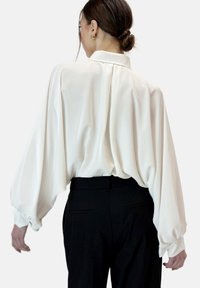 Blusa blanca, fluida, con un cuello alto y mangas abullonadas recogidas, llevada sobre pantalones negros. Tela suave con un ligero brillo.