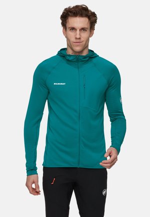 Homme portant une veste à capuche zippée teal avec une poche poitrine et un pantalon noir avec une poche zippée, se tenant devant un fond uni.