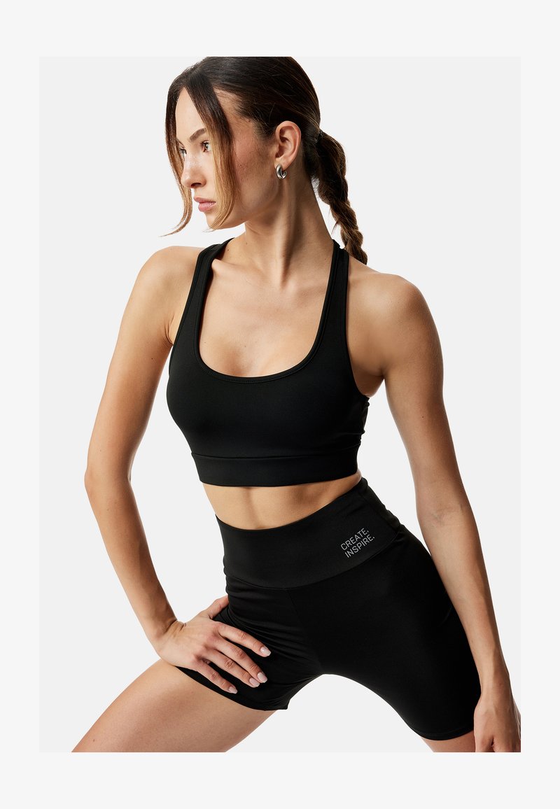 Kobieta w czarnym sportowym staniku i wysokich szortach z napisem „CREATE INSPIRE”, pozująca z ręką na biodrze, włosy związane w pleciony kucyk.