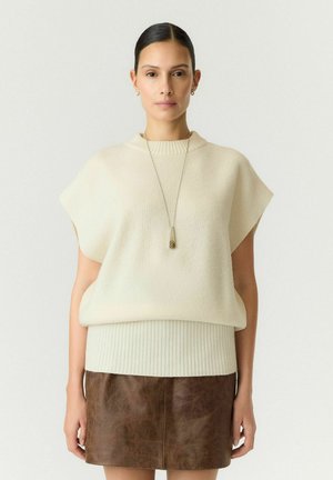 Femme aux cheveux foncés portant un pull sans manches beige et une jupe en cuir marron, ainsi qu'un long collier pendentif, debout devant un fond uni.
