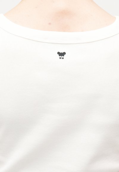 Gros plan sur l'encolure d'une chemise blanche côtelée montrant un petit logo papillon noir avec les lettres "W M" en dessous.
