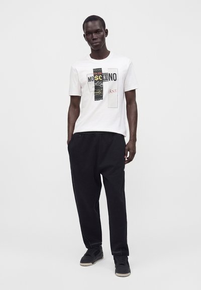 Άνδρας που φοράει λευκό T-shirt με μοτίβο Moschino, μαύρο παντελόνι και μαύρα αθλητικά παπούτσια, στέκεται μπροστά σε απλό φωτεινό φόντο.