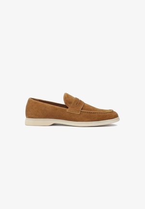 Mocassin en daim marron avec un bout arrondi, des détails surpiqués et une petite bride décorative. Il présente une semelle en caoutchouc blanche contrastante.