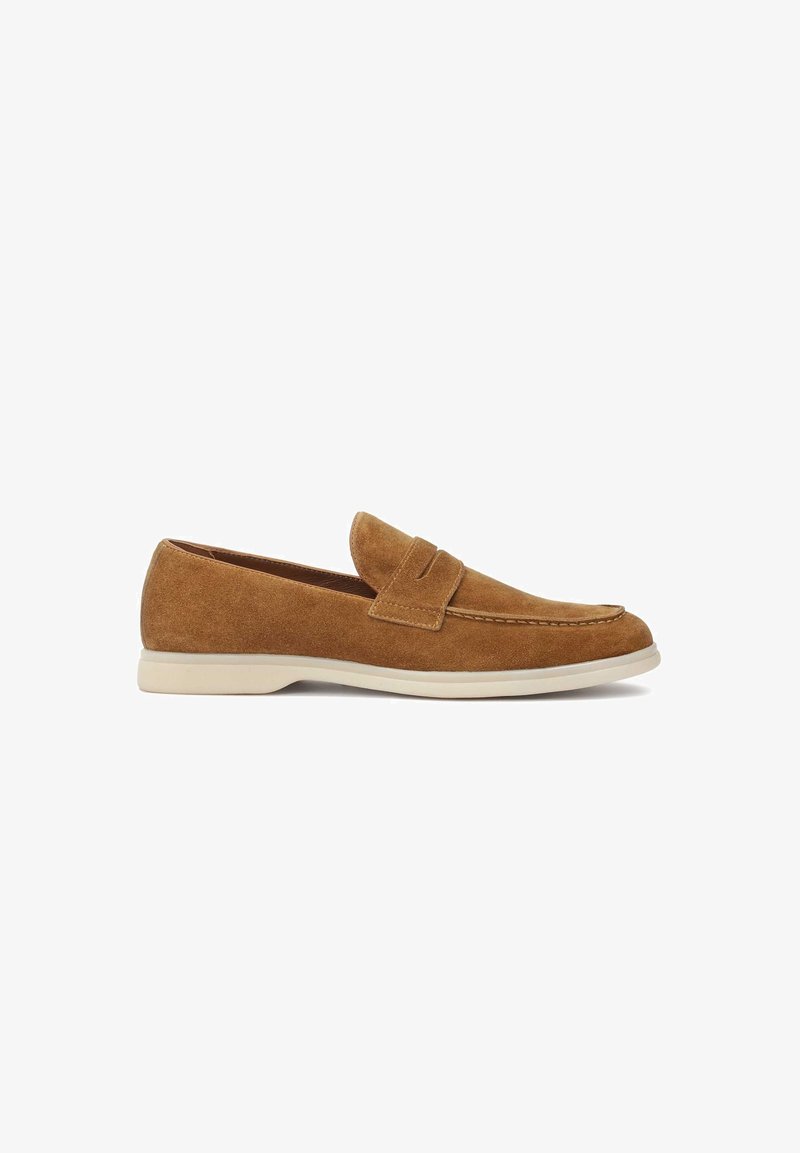 Mocassin en daim marron avec un bout arrondi, des détails surpiqués et une petite bride décorative. Il présente une semelle en caoutchouc blanche contrastante.