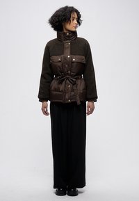 Hnědá nadýchaná bunda s texturovaným fleecovým horním dílem, stahovací šňůrkou v pase, patentními knoflíky a velkými předními kapsami. Kombinována s černými kalhotami s širokými nohavicemi.