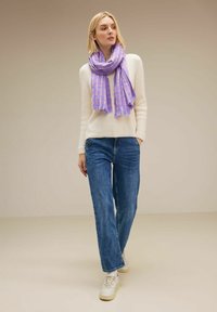 Sciarpa di un leggero viola con un design a motivo, indossata sopra un maglione crema e jeans blu. La sciarpa presenta estremità frangiate e una consistenza morbida.