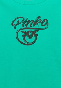 Tyrkysové tričko s kulatým výstřihem a velkým černým grafickým nápisem "Pinko" a dvěma stylizovanými ptáky pod nápisem.