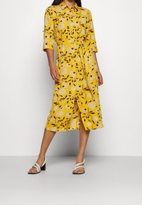 Gul blommönstrad klänning med krage, trekvartsärmar, midjebälte och en slits framtill. Matchad med vita klacksandaler.