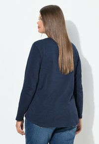 Marineblaues Langarm-T-Shirt mit abgerundetem Saum und weicher Textur, kombiniert mit blauen Jeans. Schlichtes Design ohne sichtbare Muster oder Akzente.