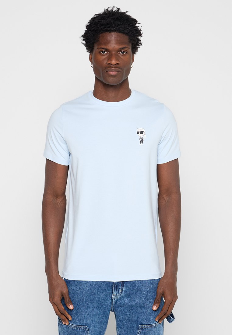 Joven con cabello corto y rizado que lleva una camiseta azul claro con un pequeño gráfico en blanco y negro en el pecho y vaqueros azules.
