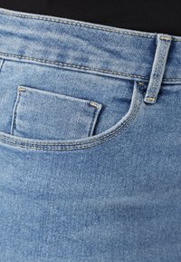 Jeans de denim azul claro com um bolso traseiro, costura detalhada e um cós. O tecido tem uma textura suave e um leve desgaste.