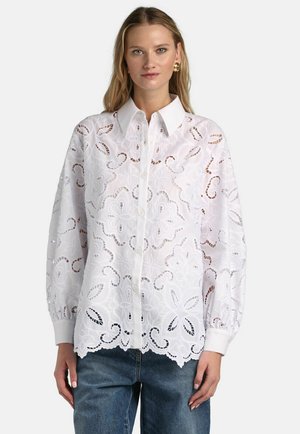 Donna che indossa una camicetta bianca con bottoni, ricamata con motivi floreali traforati, maniche lunghe a sbuffo e colletto appuntito, abbinata a jeans blu.