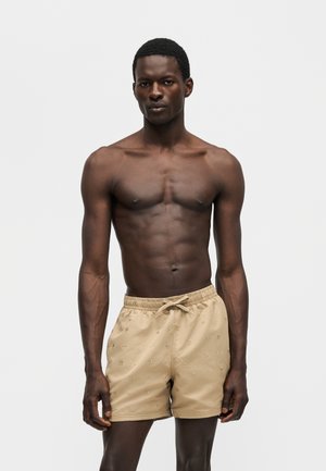 Męski, muskularny mężczyzna o ciemnej karnacji, ubrany w beżowe szorty z troczkami, stojący na jednolitym jasnoszarym tle z neutralnym wyrazem twarzy.