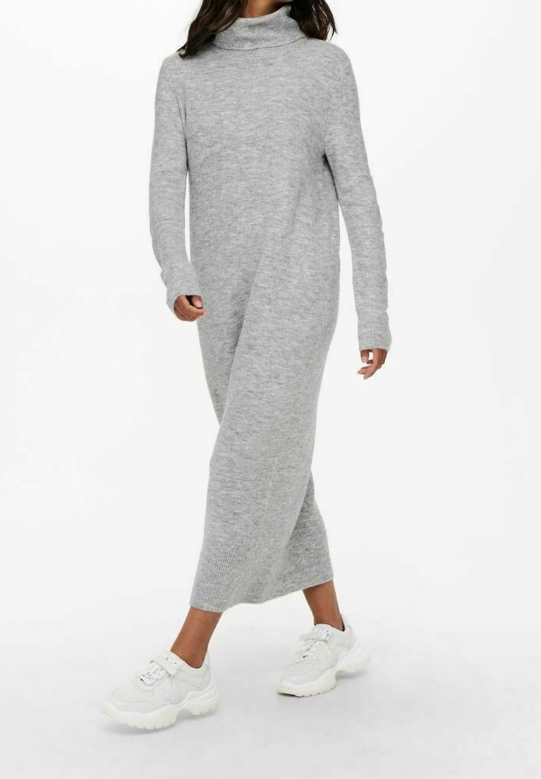 Femme portant une longue robe-pull grise à col roulé et des baskets blanches épaisses, marchant avec une posture détendue sur un fond blanc.