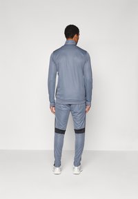 Grijze sportieve trainingsset met een ritskraagjack en tapered broek met zwarte accenten. Soepele stof met lange mouwen en elastische enkels.