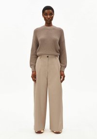 Vrouw staand, gekleed in een taupekleurige gebreide trui, ingestopt in een high-waisted beige broek met wijde pijpen, en bruine en gouden schoenen.