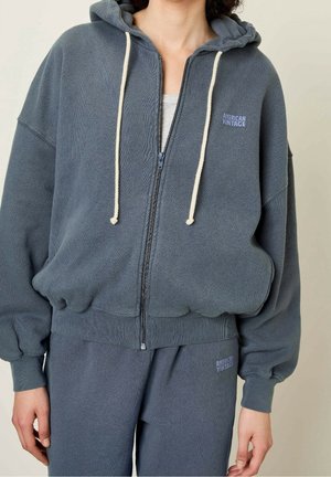 Mannequin portant un sweat à capuche gris zippé et un pantalon de survêtement assorti, tous deux avec le logo "American Vintage" sur la poitrine et la cuisse.