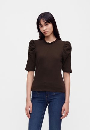 Jeune femme aux longs cheveux foncés porte un haut marron à manches bouffantes et un jean bleu, debout devant un fond clair uni.