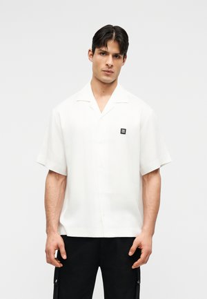 Tânăr bărbat purtând o cămașă albă cu mâneci scurte, cu un mic logo negru pe piept, și pantaloni negri, stând în fața unui fundal simplu.