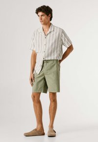 Άνδρας που φοράει κοντομάνικο πουκάμισο με ρίγες, χακί σορτς και καφέ παπούτσια τύπου slip-on, στέκεται μπροστά σε απλό φόντο.