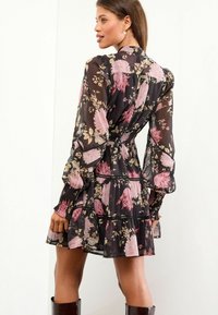Rochie neagră cu flori, cu mâneci lungi și transparente și talie strânsă. Prezintă modele florale roz și crem și volane în straturi la tiv.