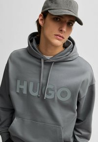 Grijze hoodie met grote "HUGO"-lettering in een lichtere tint, trekkoorden en een kangoeroezak. De stof heeft een gladde textuur.