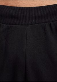 Schwarze Jogginghosen verfügen über einen gerippten Bund mit einer glatten Textur, tiefen Falten und einem schlichten Design, das auf Komfort und Trageease fokussiert ist.